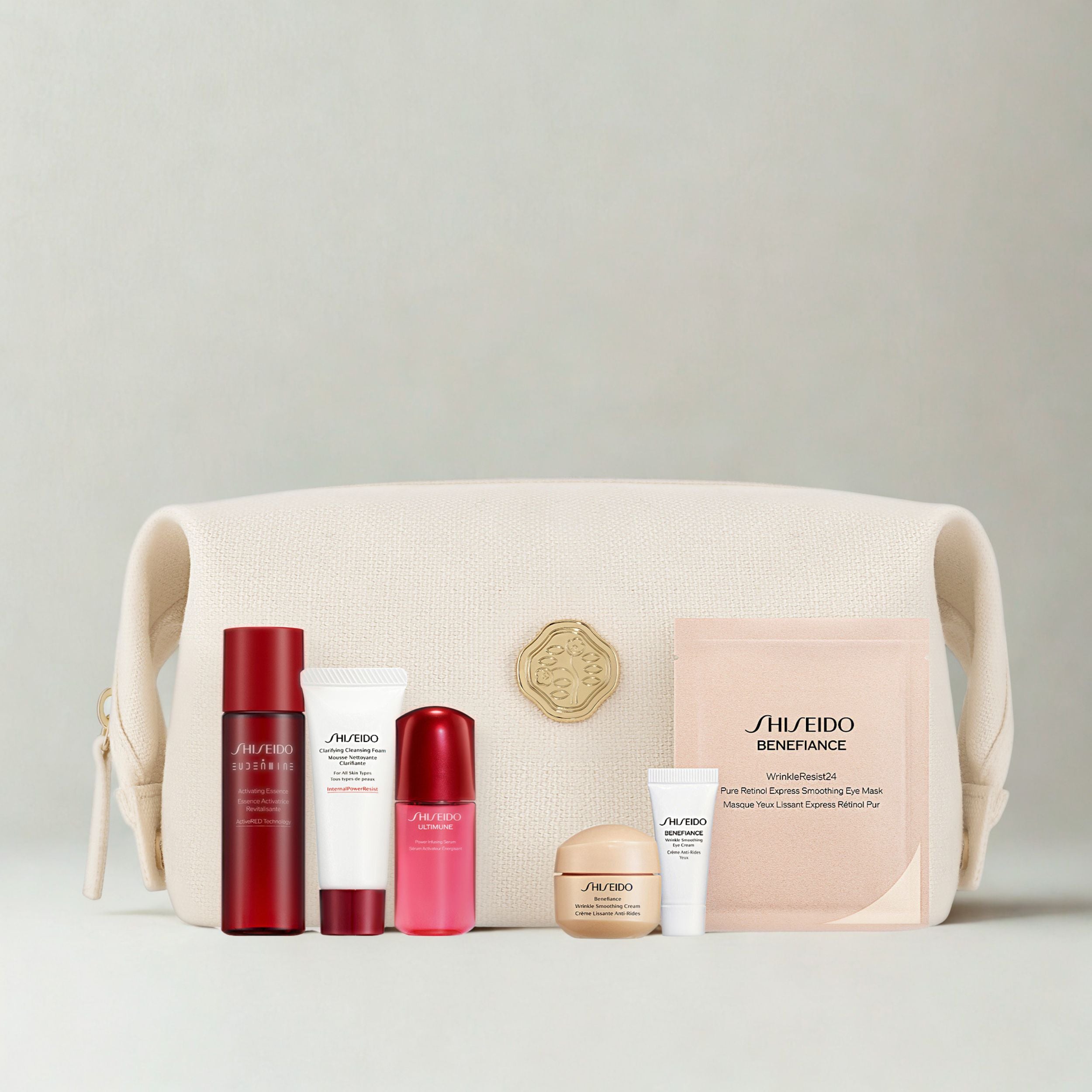 A Shiseido gift set arranged in front of a cream-colored canvas pouch. The set includes a red bottle, a white tube, a pink bottle, a gold jar, a white tube, and a pink packet, all on a light gray background.