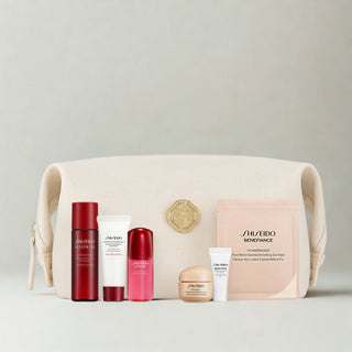 A Shiseido gift set arranged in front of a cream-colored canvas pouch. The set includes a red bottle, a white tube, a pink bottle, a gold jar, a white tube, and a pink packet, all on a light gray background.