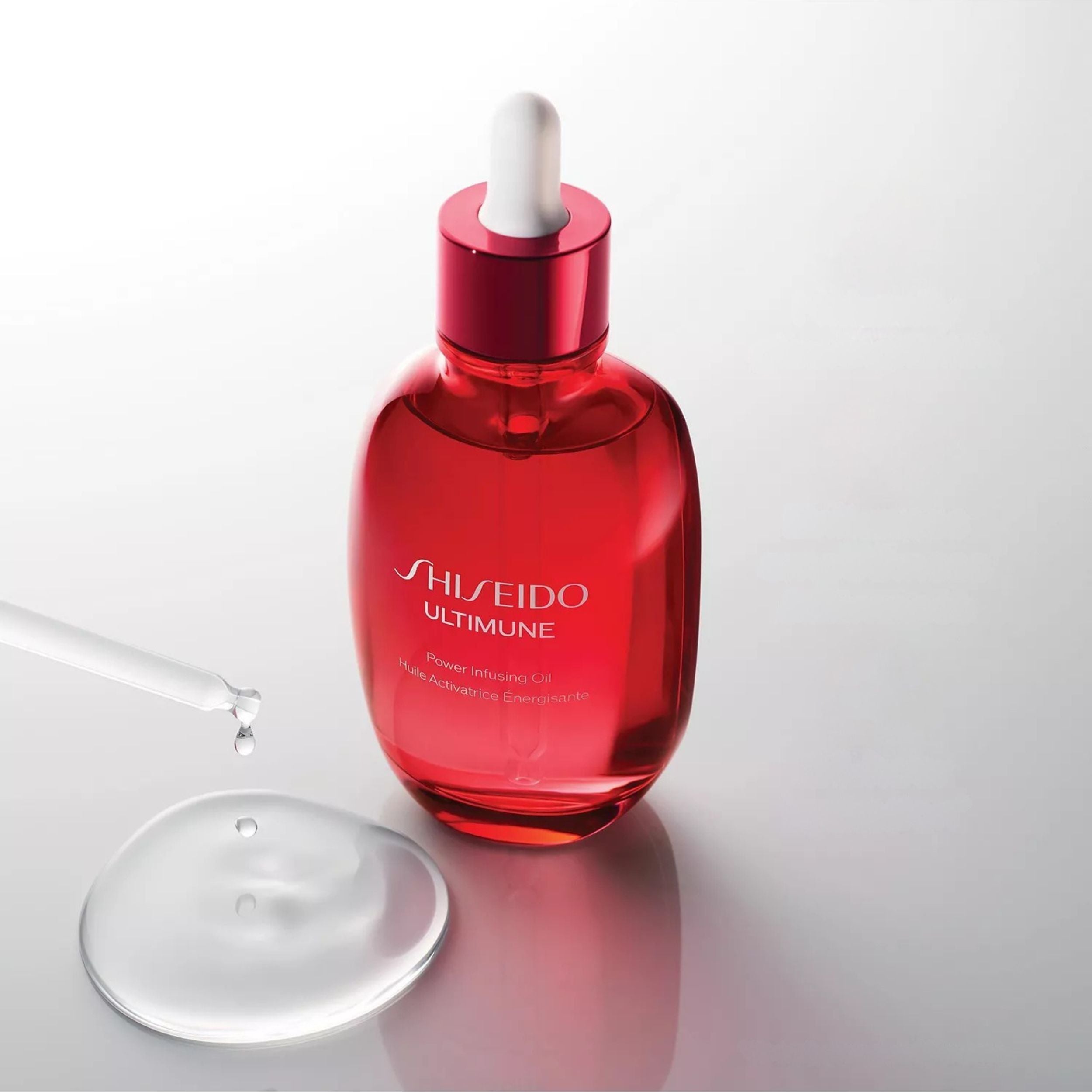 Shiseido Ultimune Power-protecting Serum in a translucent red glass dropper bottle, shown with a glass pipette and a clear droplet on a light gray reflective surface.